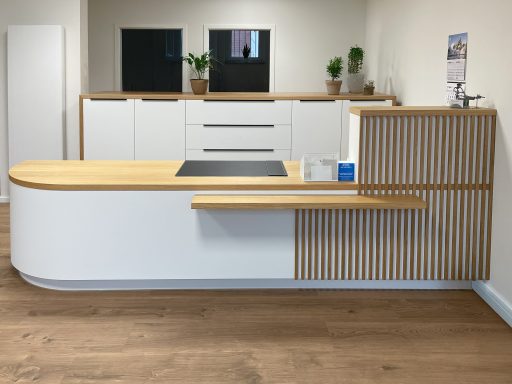 Weiße Empfangstresen mit abgerundeter Holzplatte und vertikalen Holzleisten. Dahinter weiße Stauraumschränke. Minimalistisches Design.