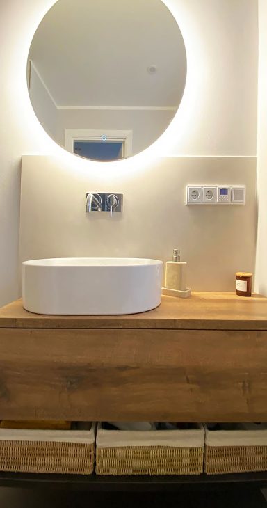Holz-Waschtisch mit rundem, hinterleuchtetem Spiegel und Aufsatzwaschbecken. Darunter Körbe für Stauraum.