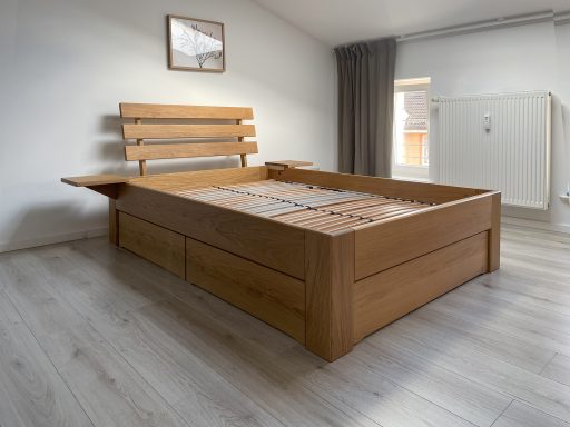 Ein massives Eichenbett mit hohem Kopfteil, integrierten Nachttischablagen und Schubladen mit viel Stauraum in einem hellen Schlafzimmer.