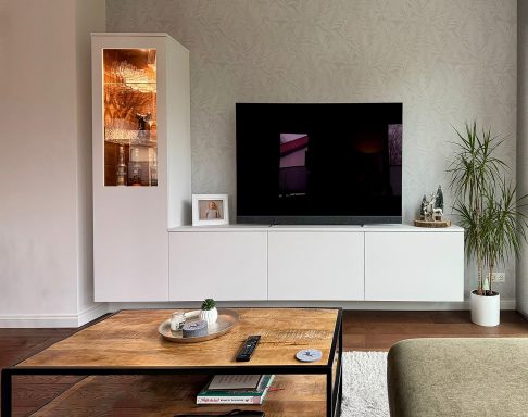 Wohnwand-Set in Weiß mit beleuchteter Glasvitrine, TV-Board und gläsernen Regalen. Rechts steht eine Zimmerpflanze. Vorn ein Holztisch.